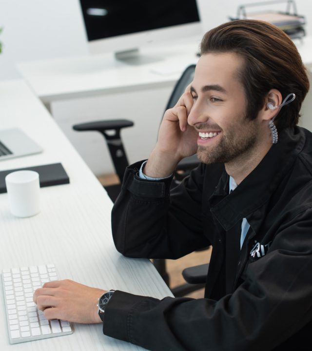 young security man in earphone smiling at workplace near computer monitor